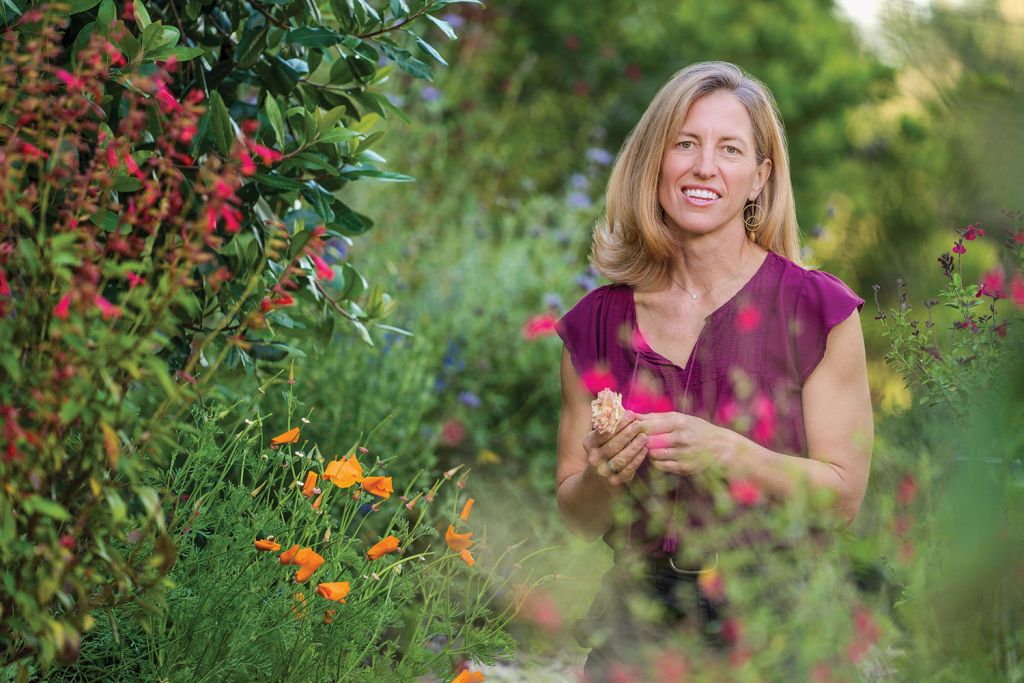 Sattely with long blond hair in purple top smiles while standing in a colorful flower garden.