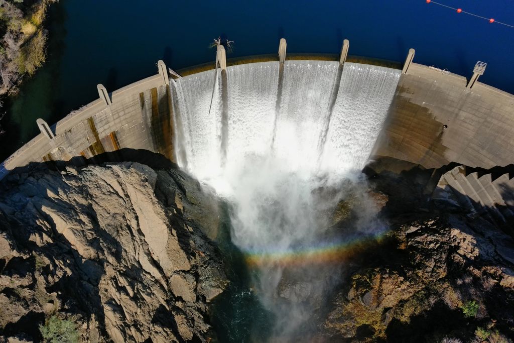 Aerial photo of water rushing over a curved concrete dam into a rocky canyon below, with a rainbow visible in the mist.