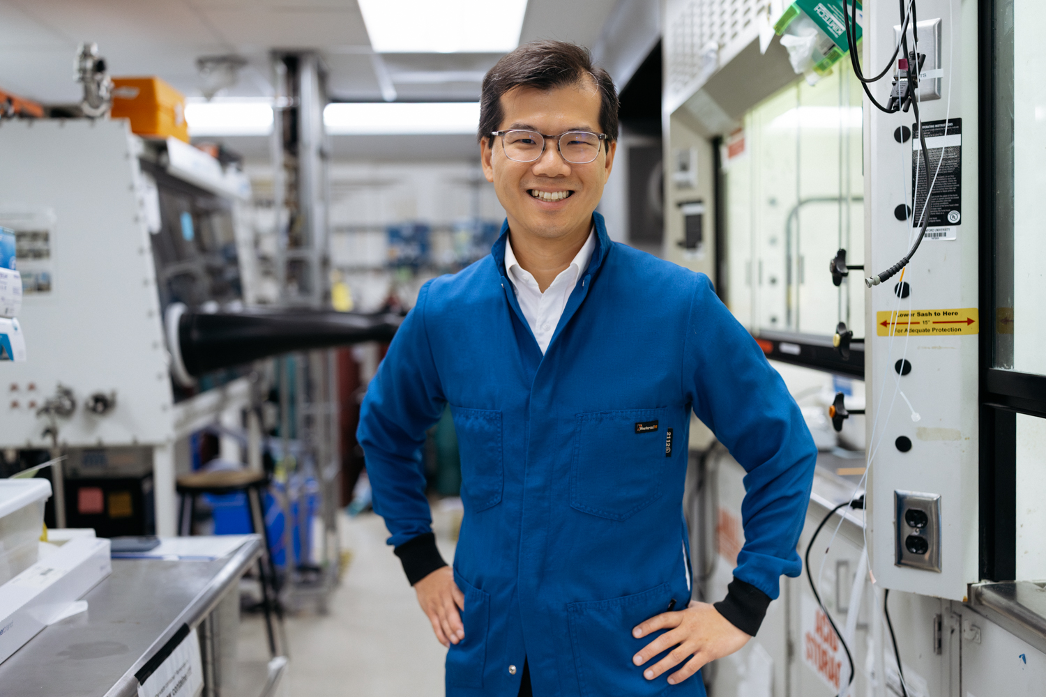 William Chueh in a blue lab coat and glasses smiles while standing among equipment in a lab.