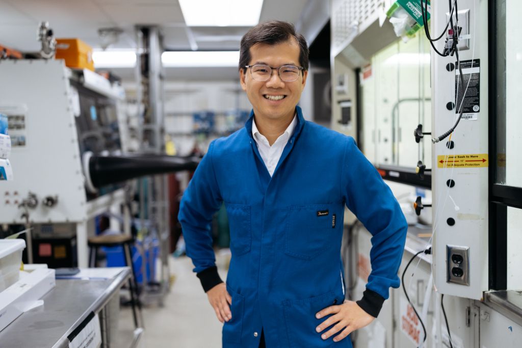 William Chueh in a blue lab coat and glasses smiles while standing among equipment in a lab.