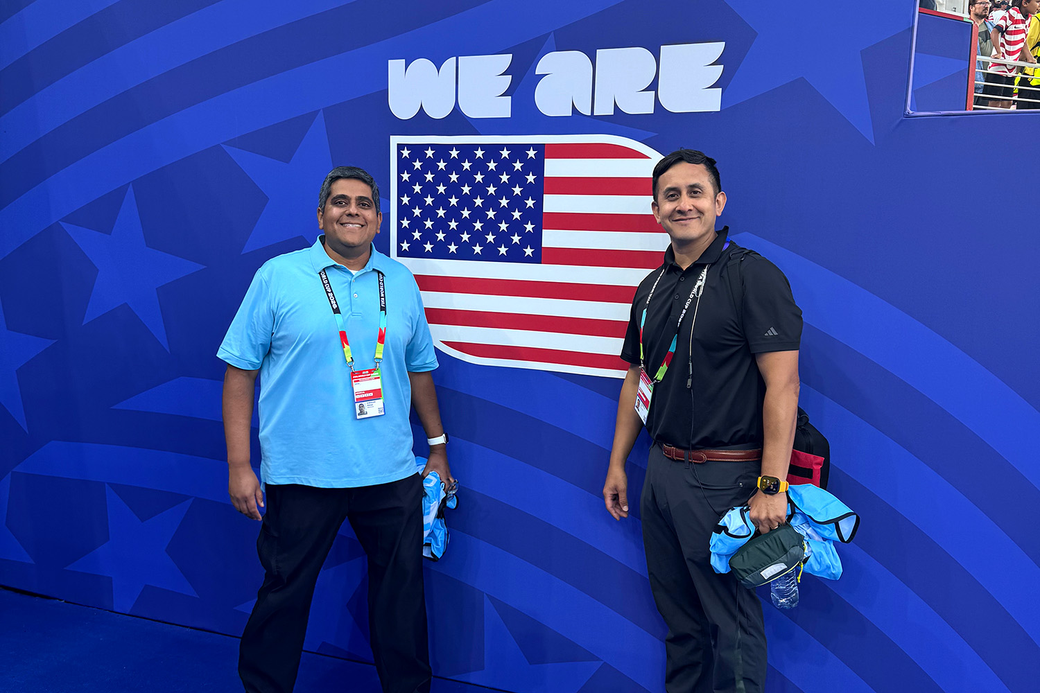 Peter D’Souza and Eli Carrillo pose in front of a blue wall showing a U.S. flag graphic at a soccer venue.