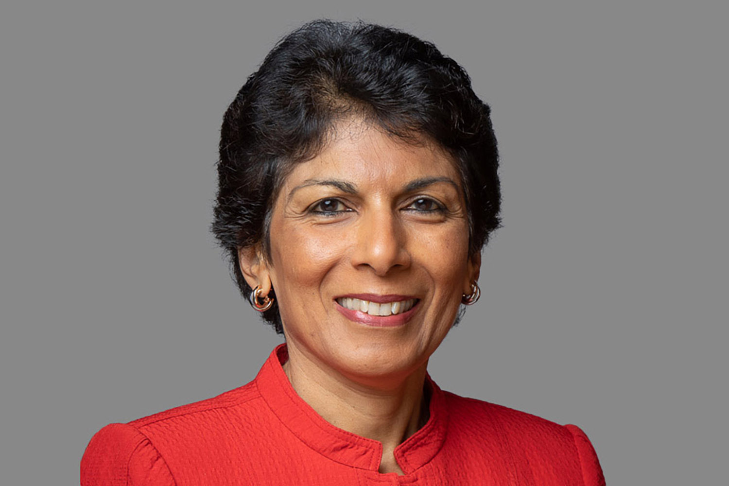 Smiling Farah Champsi wearing a red jacket poses against a gray background.