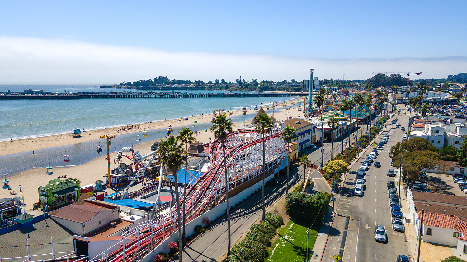 Aerial view of Santa Cruz boardwalk with a roller coaster and palm trees, alongside a sandy beach and ocean waves.