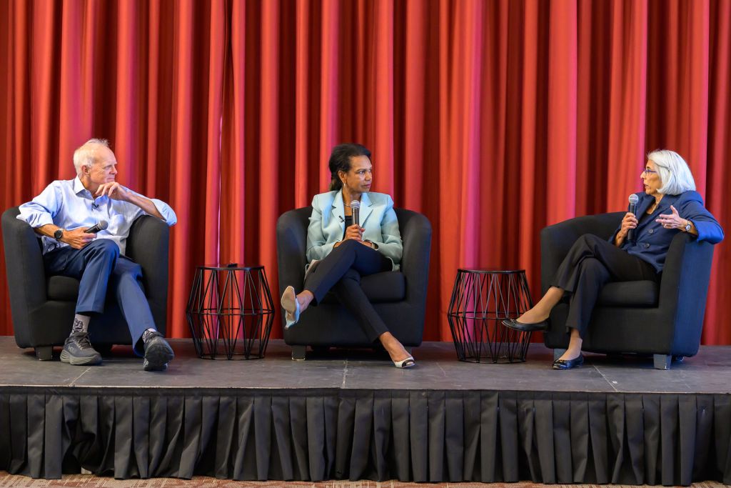 Three panelists seated in armchairs on a stage with a red curtain backdrop, holding microphones during a discussion.