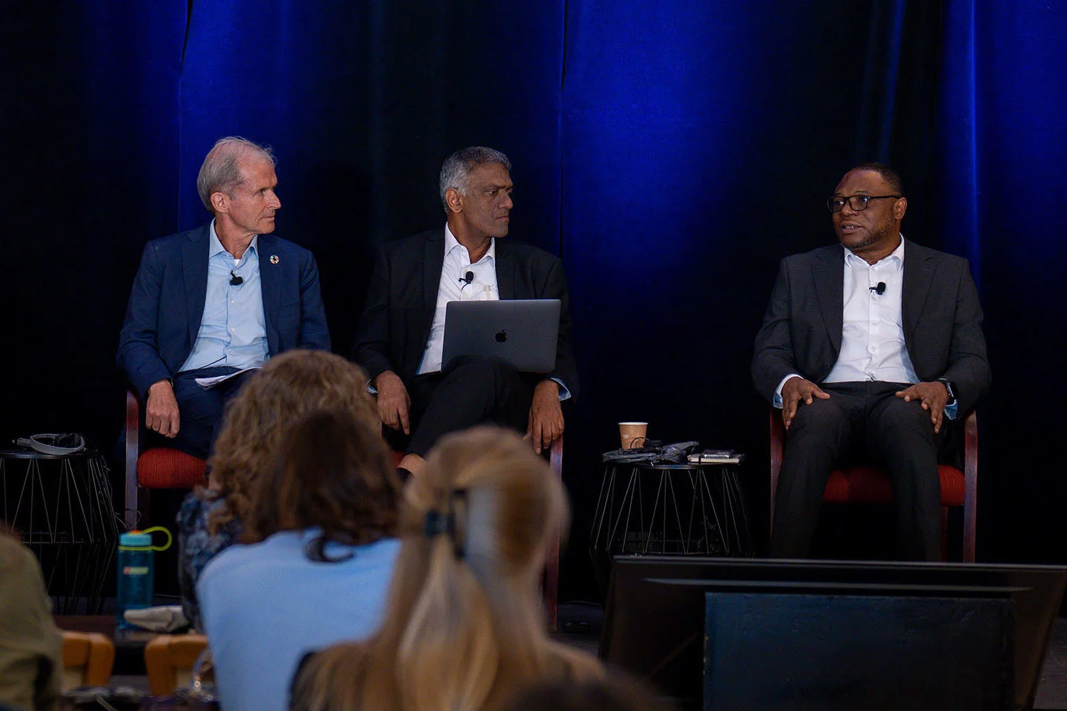 Three men in suits sit on stage during a panel discussion, with an audience visible in the foreground.