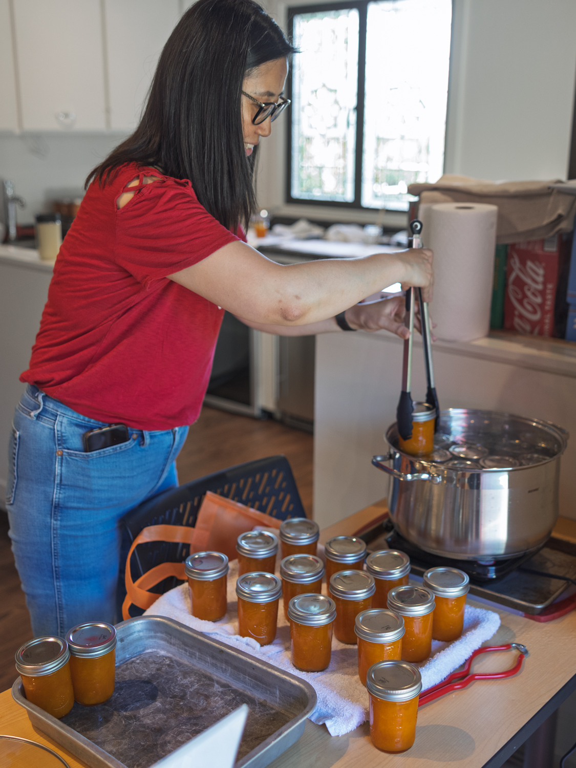 Woman using a jar lifter to remove hot jam jars from a canning pot in a home kitchen.