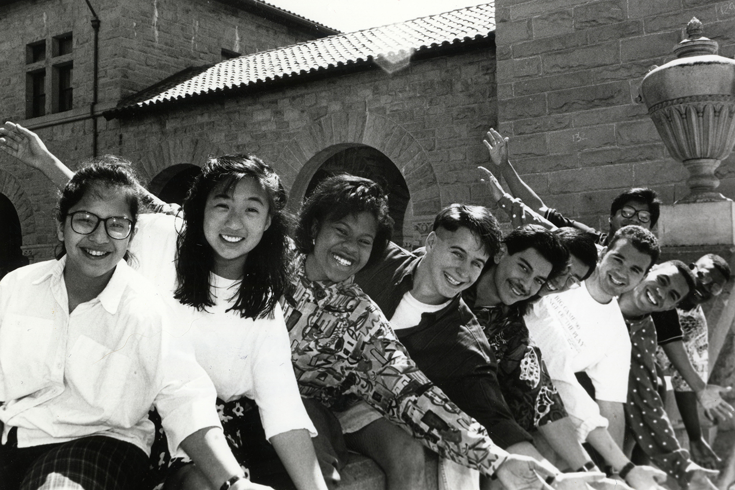 Smiling students leaning in a row outside with Quad in the background.