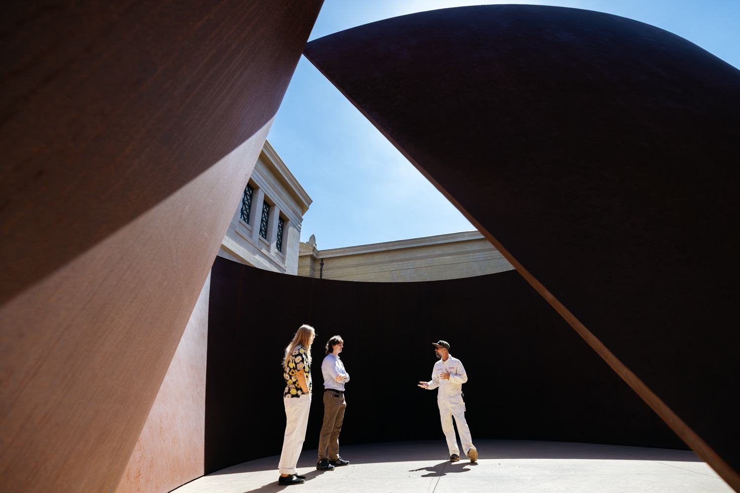 Two people plus art ranger Mark Shunney stand talking inside a towering curved steel sculpture that frames a slice of blue sky above them.