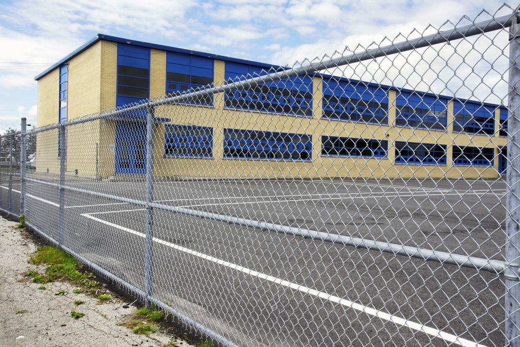 A school building with blue windows is seen behind a chain-link fence.