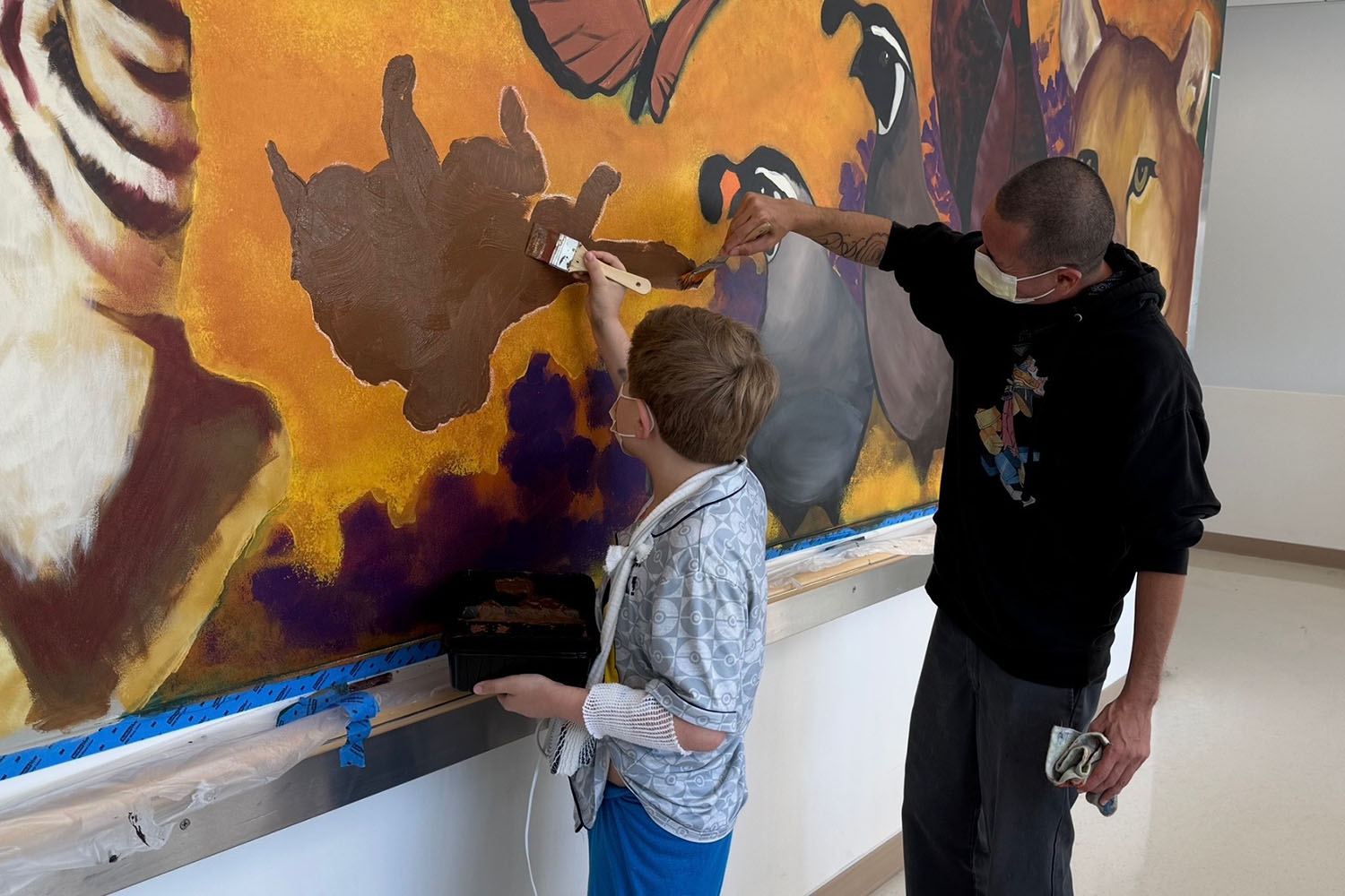 A man and a young hospital panttient in masks paint part of a wildlife mural together.