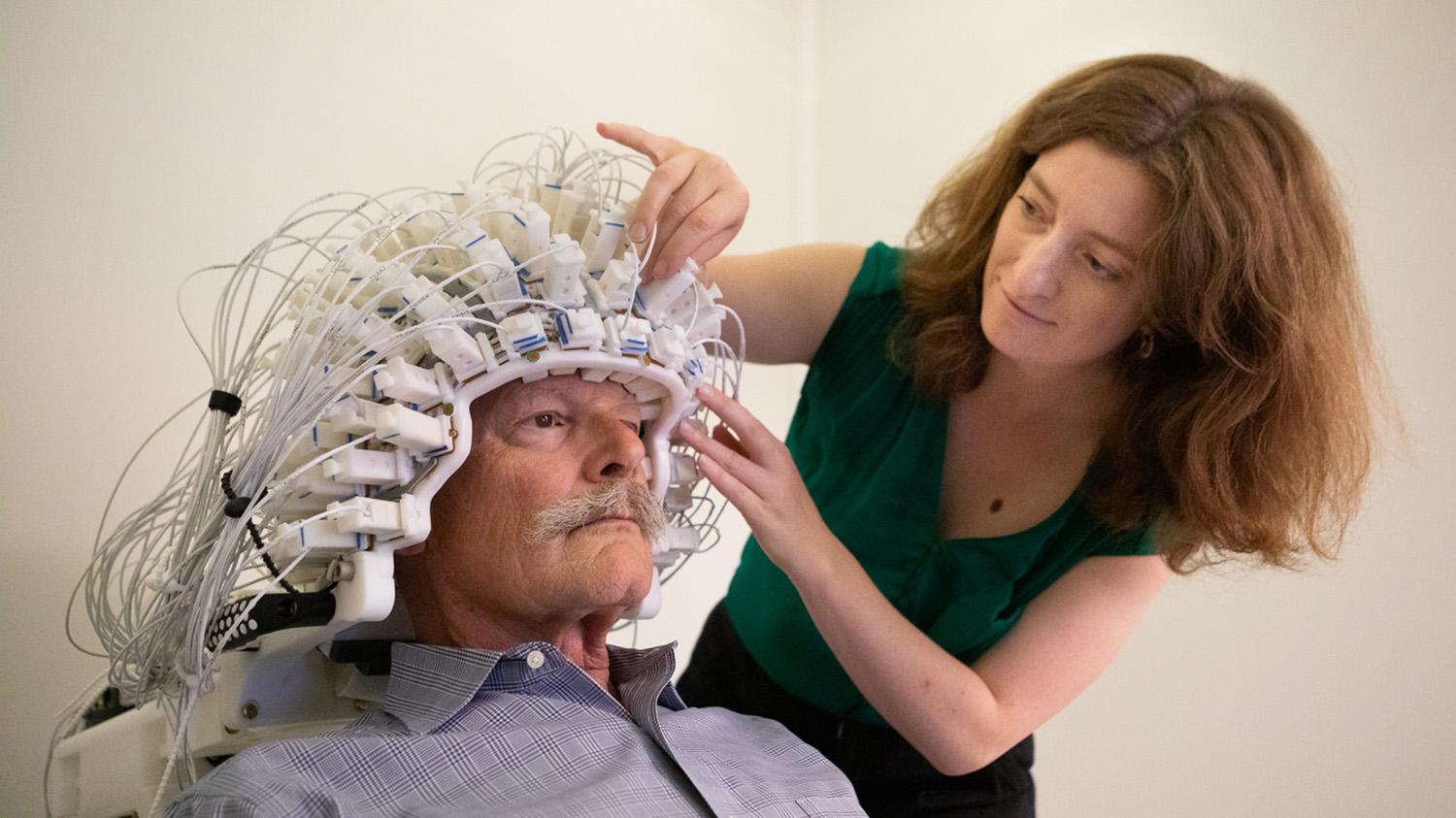 A researcher fits a wired sensor cap used for brain imaging onto a seated study participant.