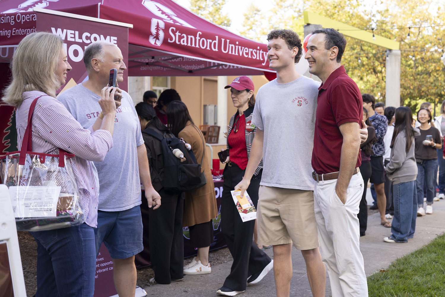A student poses with President Jonathan Levin near a Stanford University welcome tent surrounded by other arriving families.