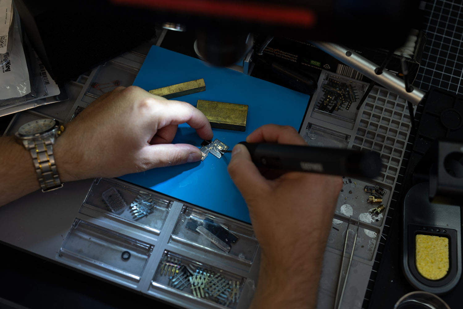 Mitchell Peterson's hands in close-up use a soldering iron and tweezers to work on a tiny circuit board over a blue mat.