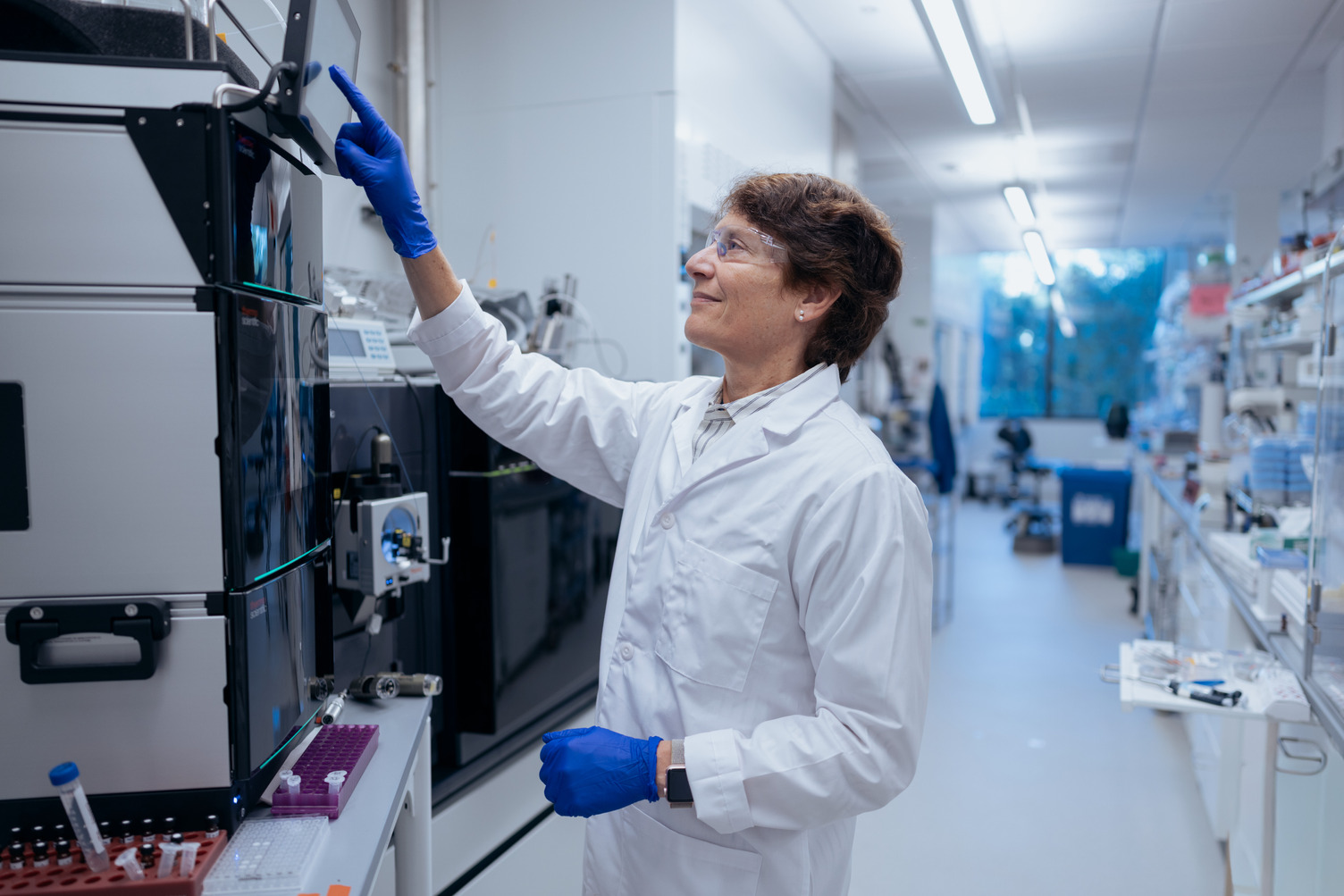 Carolyn Bertozzi in a white lab coat, safety glasses, and blue gloves reaches up to touch the screen of an analytical instrument in a lab.