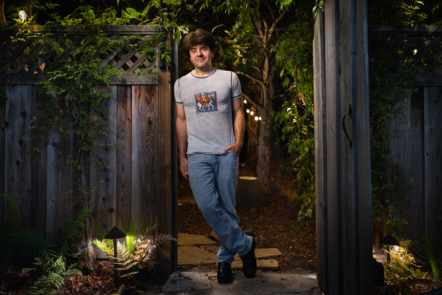 Karl Deisseroth leans against an open wooden gate at night, with a softly lit garden path and trees behind him.