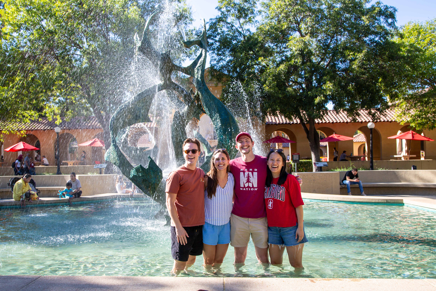 Four friends stand arm in arm in a pool of water, smiling as water sprays from White Memorial Fountain behind them.