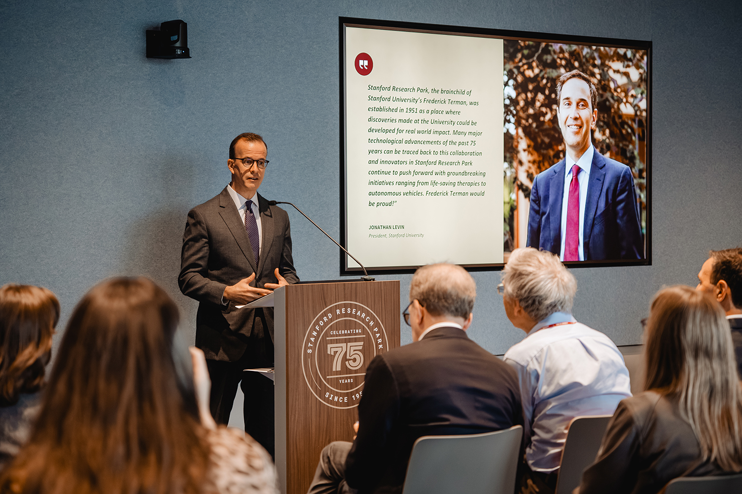 Marc Berman addresses an audience from a 75th-anniversary podium beside a screen displaying a quote from Stanford President Jonathan Levin.