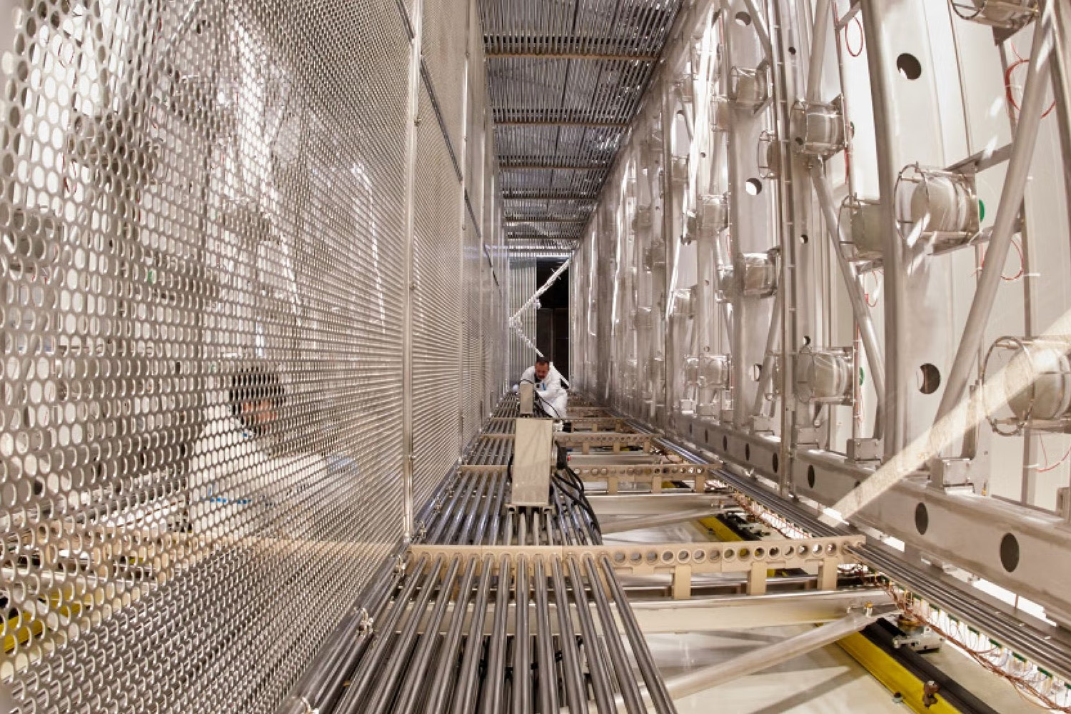 Technician in a protective suit works inside a long metal detector chamber lined with perforated panels and rows of equipment.