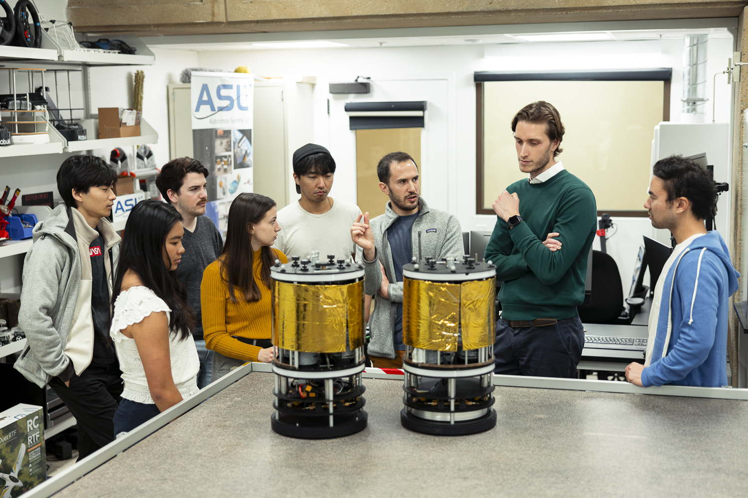 Eight people in discussion by two pieces of the robotic free flyer platform, which are cylindrical objects on top of a granite surface