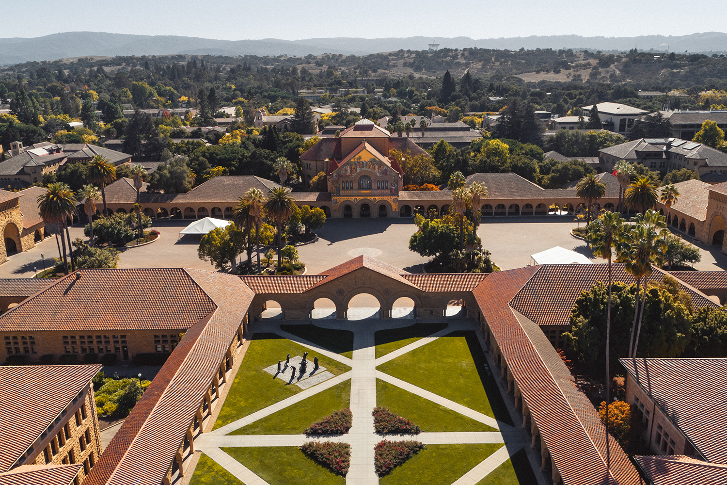 Stanford's Memorial Church, an empty Main Quad, and the grassy Memorial Court viewed from above. The rest of campus sprawls behind, all the way to the tree-lined hills.