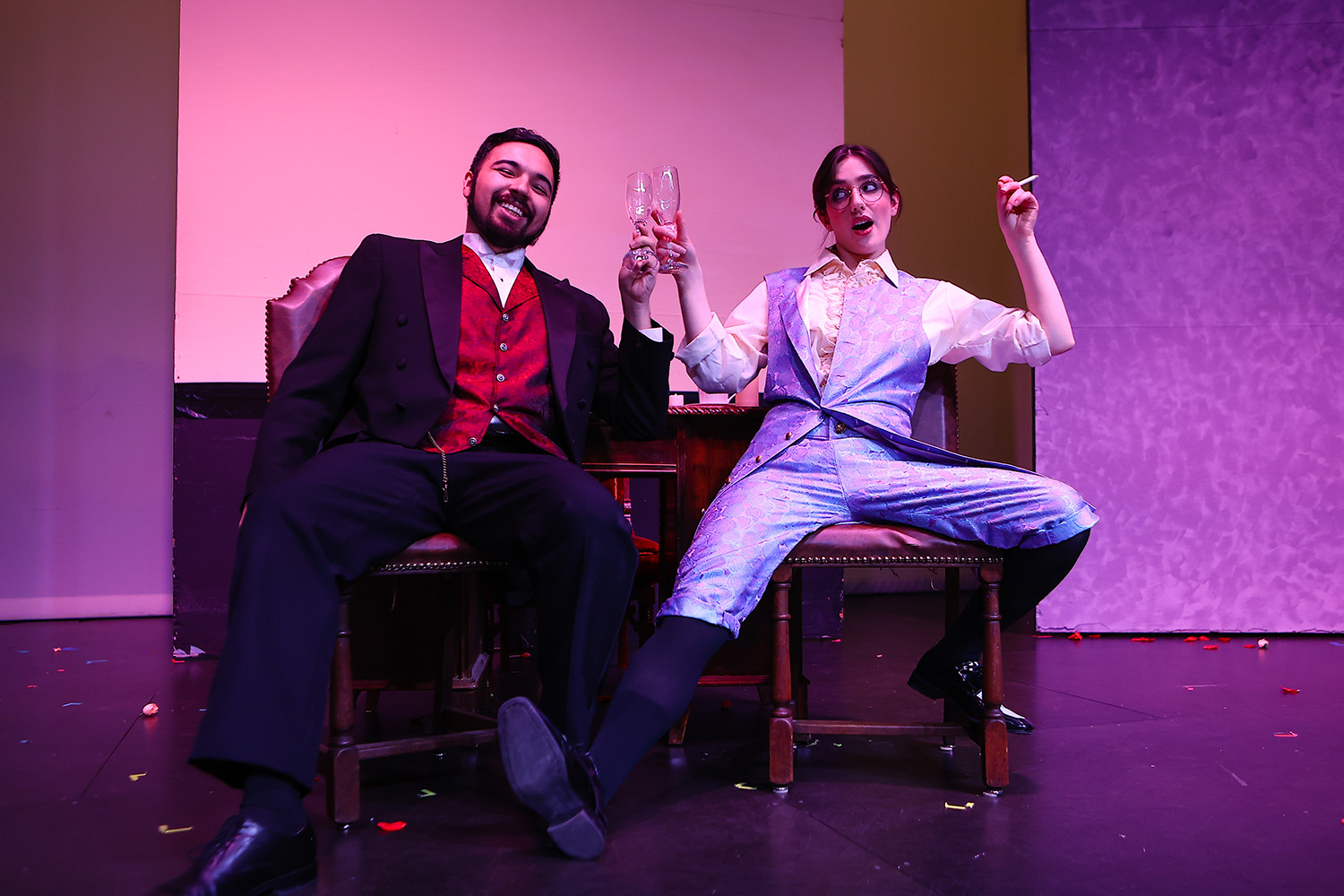 Sarah slouches jauntily in a chair facing the audience. She wears a purple-pattered vest and matching knickers and is clinking glasses with a male actor in a dark suit and a red vest. She holds a cigarette in her other hand.