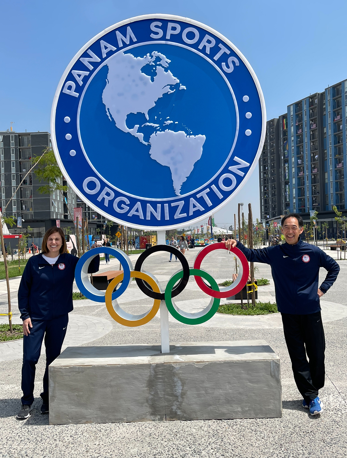 Dr. Steven Isono is pictured standing to the right and leaning with one arm on the Pan Am Sports Organization logo, with Dr. Mimi Raleigh standing on the opposite side.