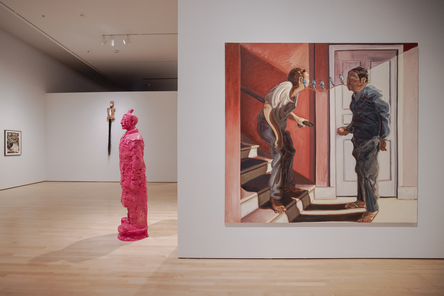 In a large gallery room, a pink wax figure of a historical Chinese soldier stands facing the room; to its right hangs a large painting of two figures facing each other in a hallway. Tiny figures walk on a wavy path between the mouth of one figure and the ear of the other.