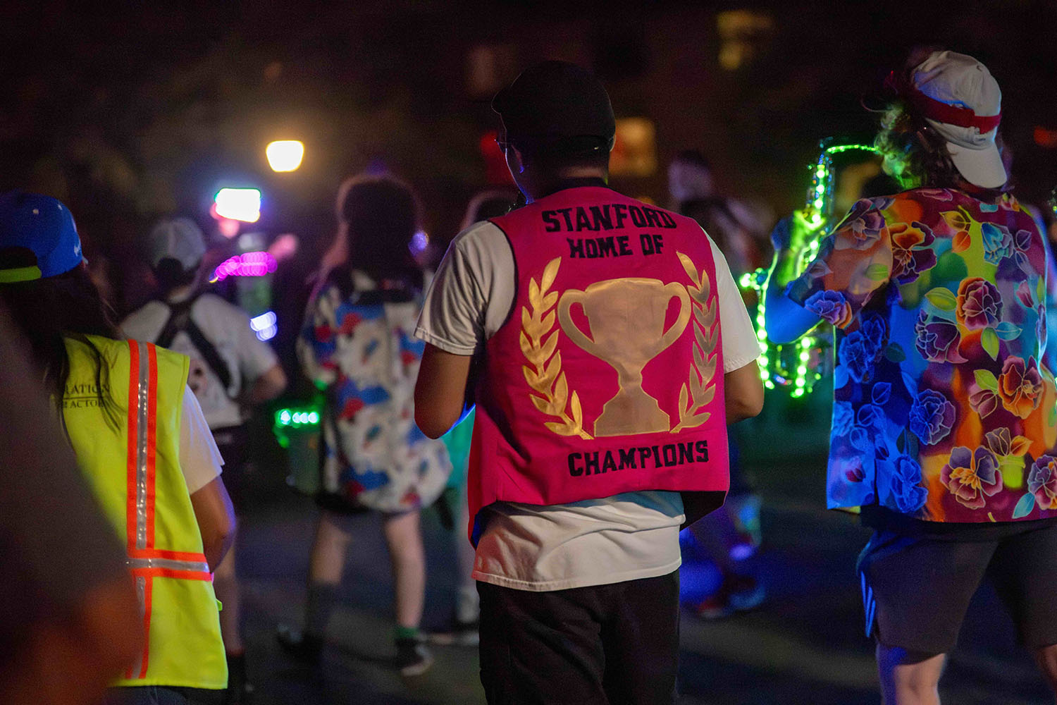 Students walking along route shot from behind. Focus is on student wearing a red vest embroidered with a trophy and the words: Stanford Home of Champions.