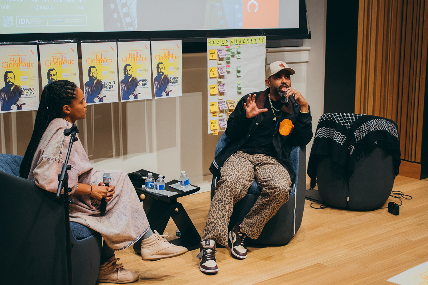 Chinaka Hodge with Daveed Diggs discussing film topics while seated in comfortable chairs, with promotional materials and a whiteboard nearby.