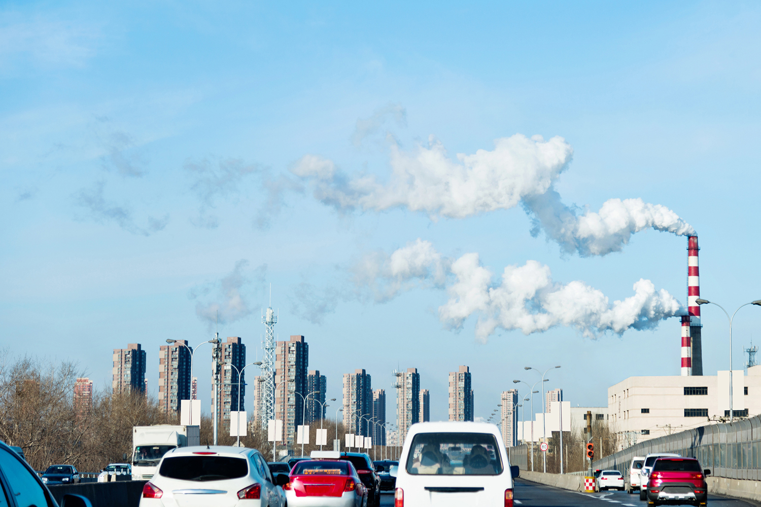 Smoke billows from a factory chimney while cars drive down a freeway, with tall buildings in the background.