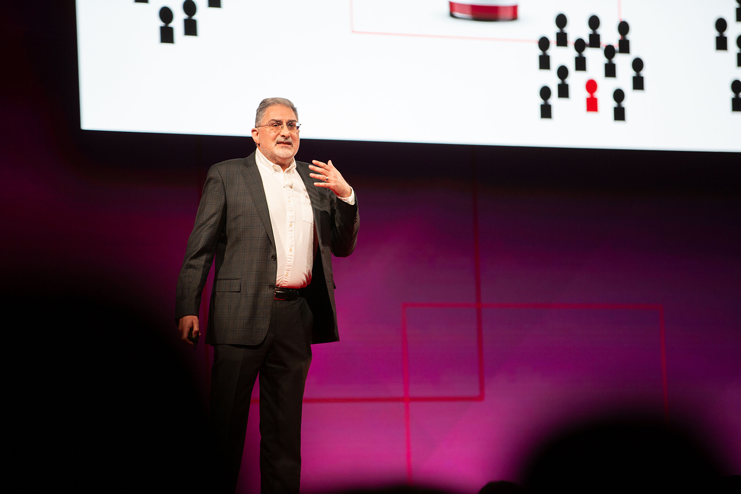 Mehran Sahami in dark suit gestures while speaking on stage with slide showing person icons.
