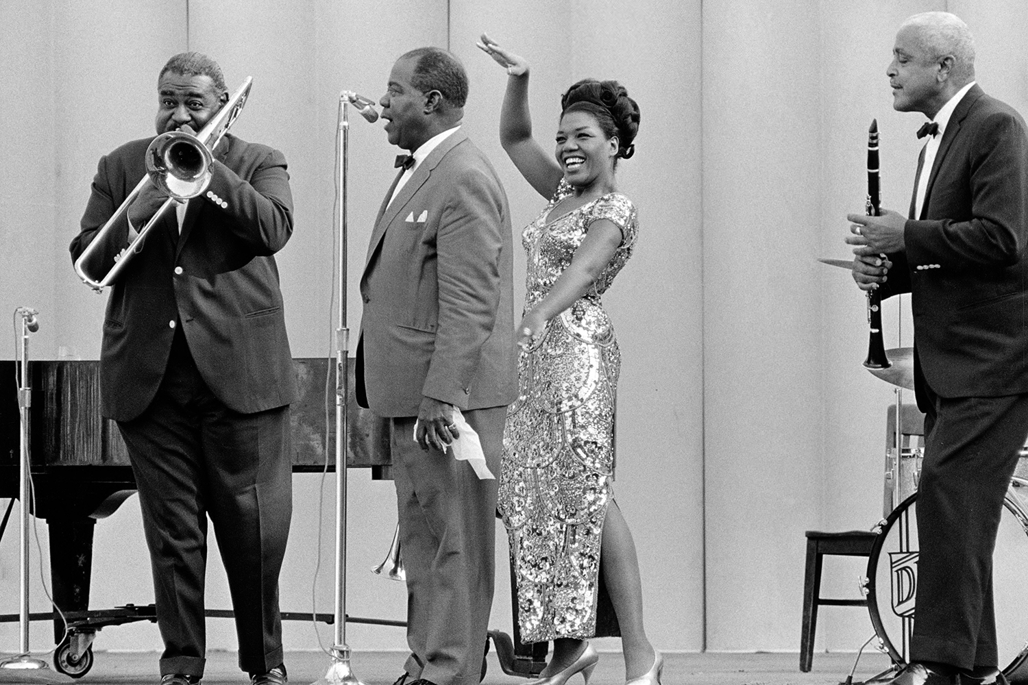 Louis Armstrong performs on stage, featuring a woman in a sparkling dress and two men playing instruments.