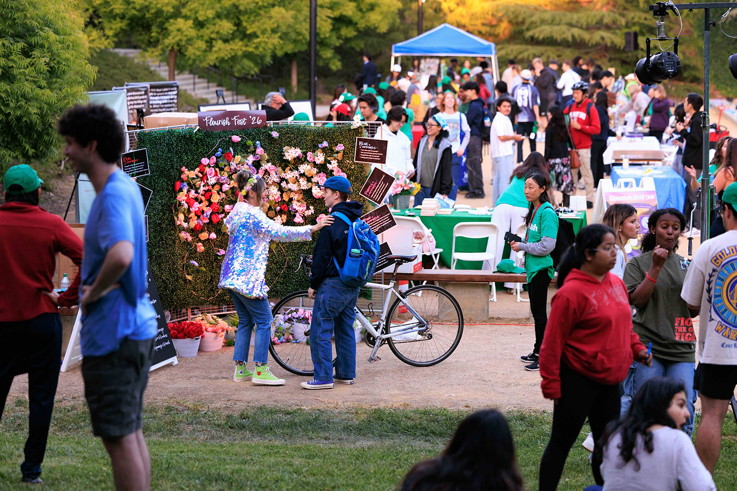 A festive outdoor scene at a community event with people interacting, a flower backdrop, and a bicycle parked nearby.