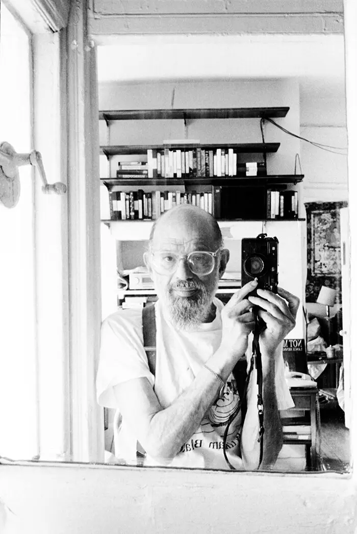 Graybeard Ginsberg takes a camera selfie in a mirror, showing bookshelves and other items in the background.