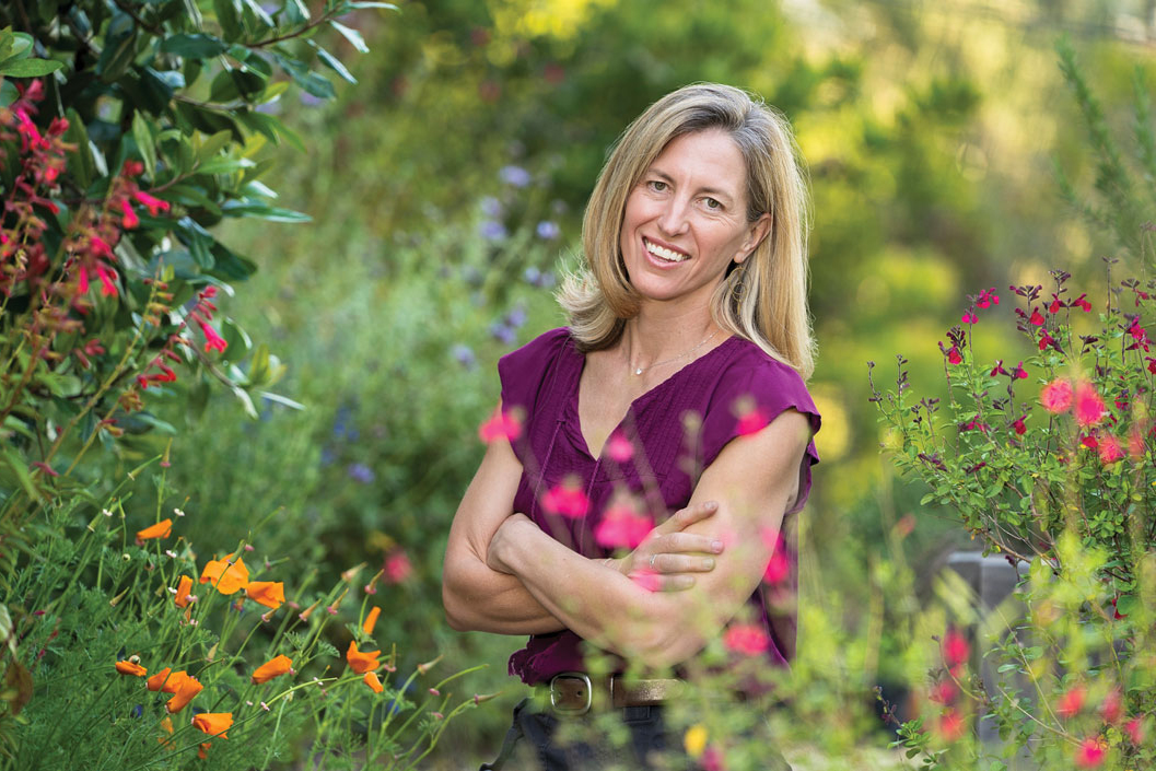 Sattely with long blond hair in purple top smiles while standing in a colorful flower garden.