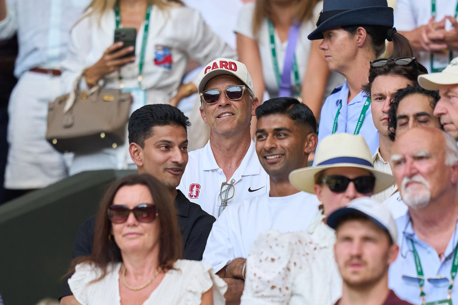 Coach Paul Goldstein in a Stanford cap and sunglasses smiles in a packed Wimbledon crowd, surrounded by other spectators.