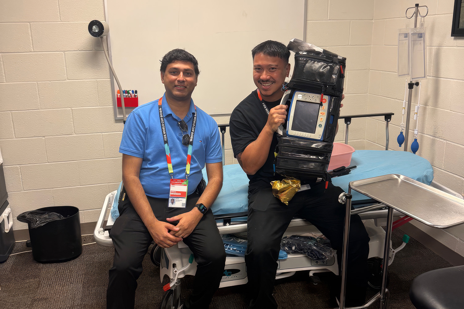 Shashank Ravi and JR Abellon sit on an exam table in a stadium treatment room; one holds up a portable heart monitor and defibrillator.