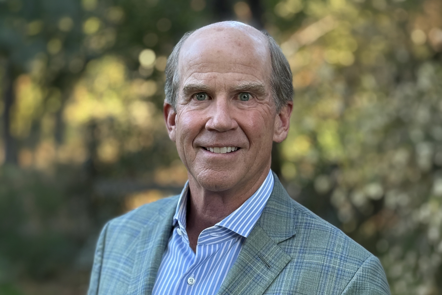 Smiling Fred Harman in a plaid blazer stands outdoors with blurred greenery in the background.