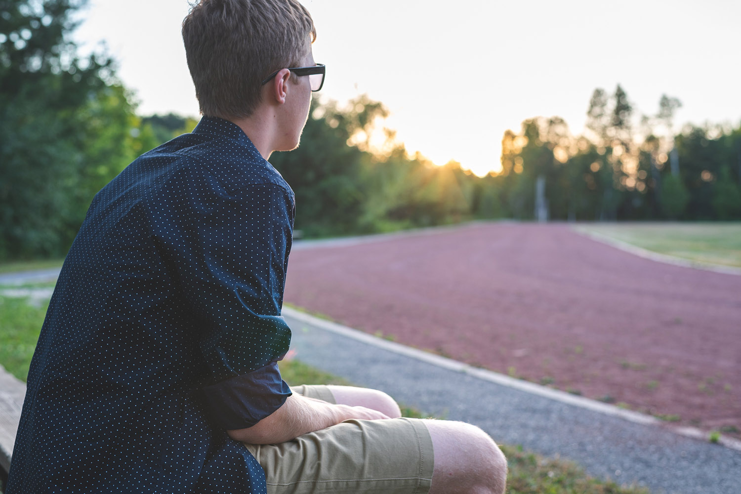 A young man sits on a bench, wearing glasses and a dotted shirt, gazing at a sunset over a running track.