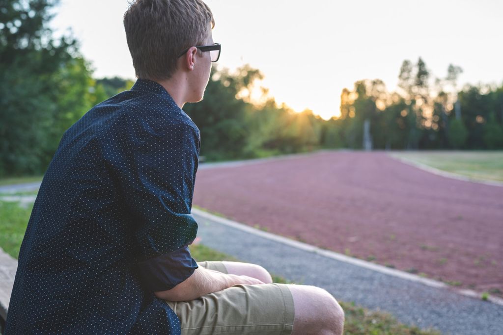 A young man sits on a bench, wearing glasses and a dotted shirt, gazing at a sunset over a running track.