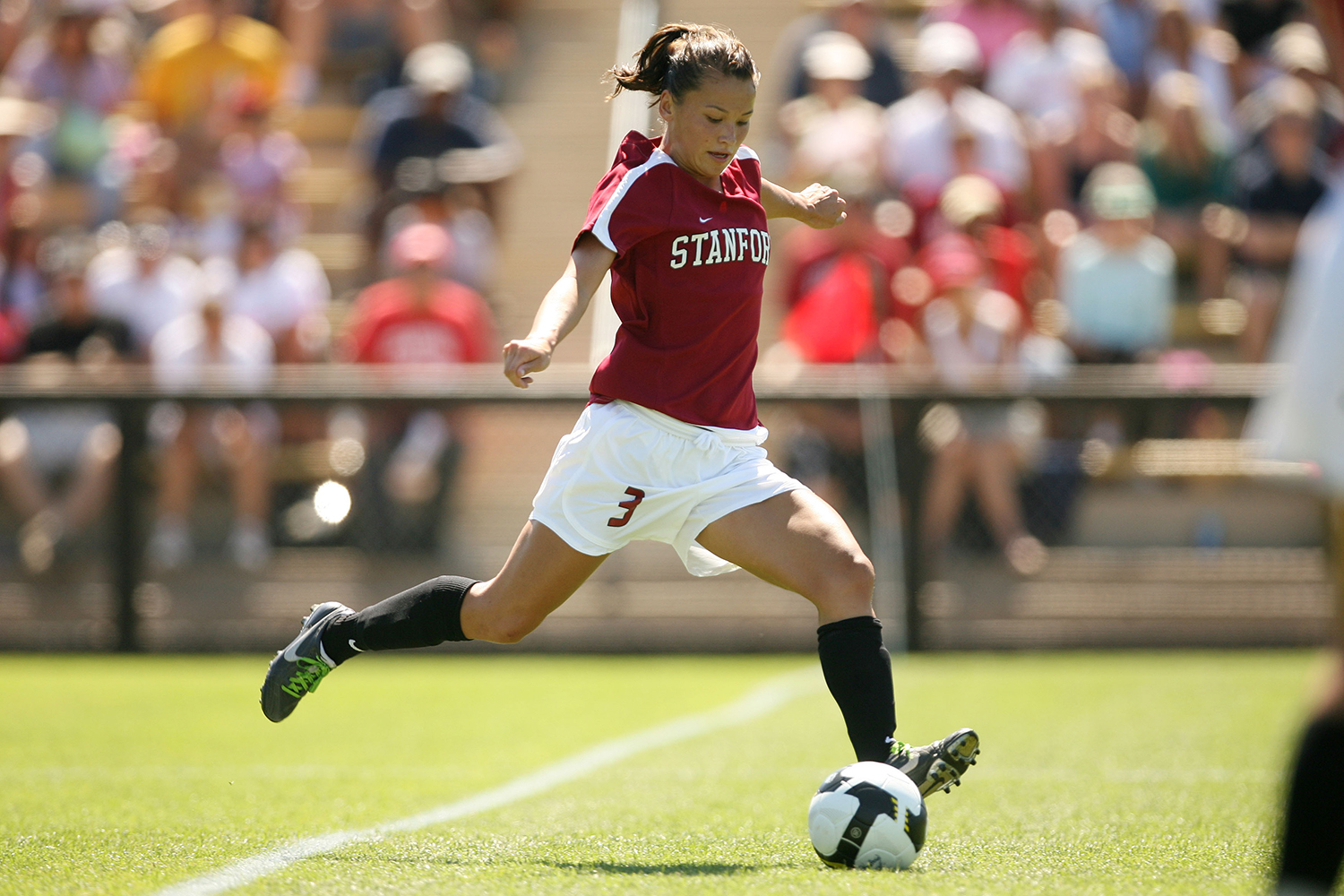 Stanford women's soccer player Ali Riley in a cardinal red jersey winds up to kick the ball during a game in front of a packed crowd.