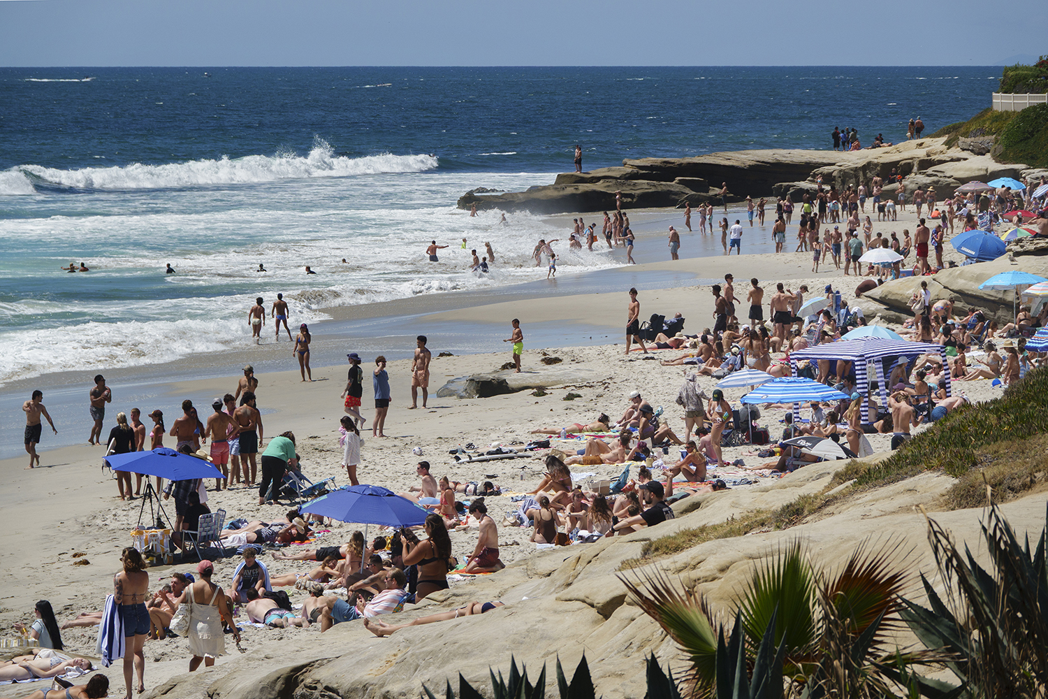 Crowds sunbathe and swim at a busy beach lined with rocks and colorful umbrellas.