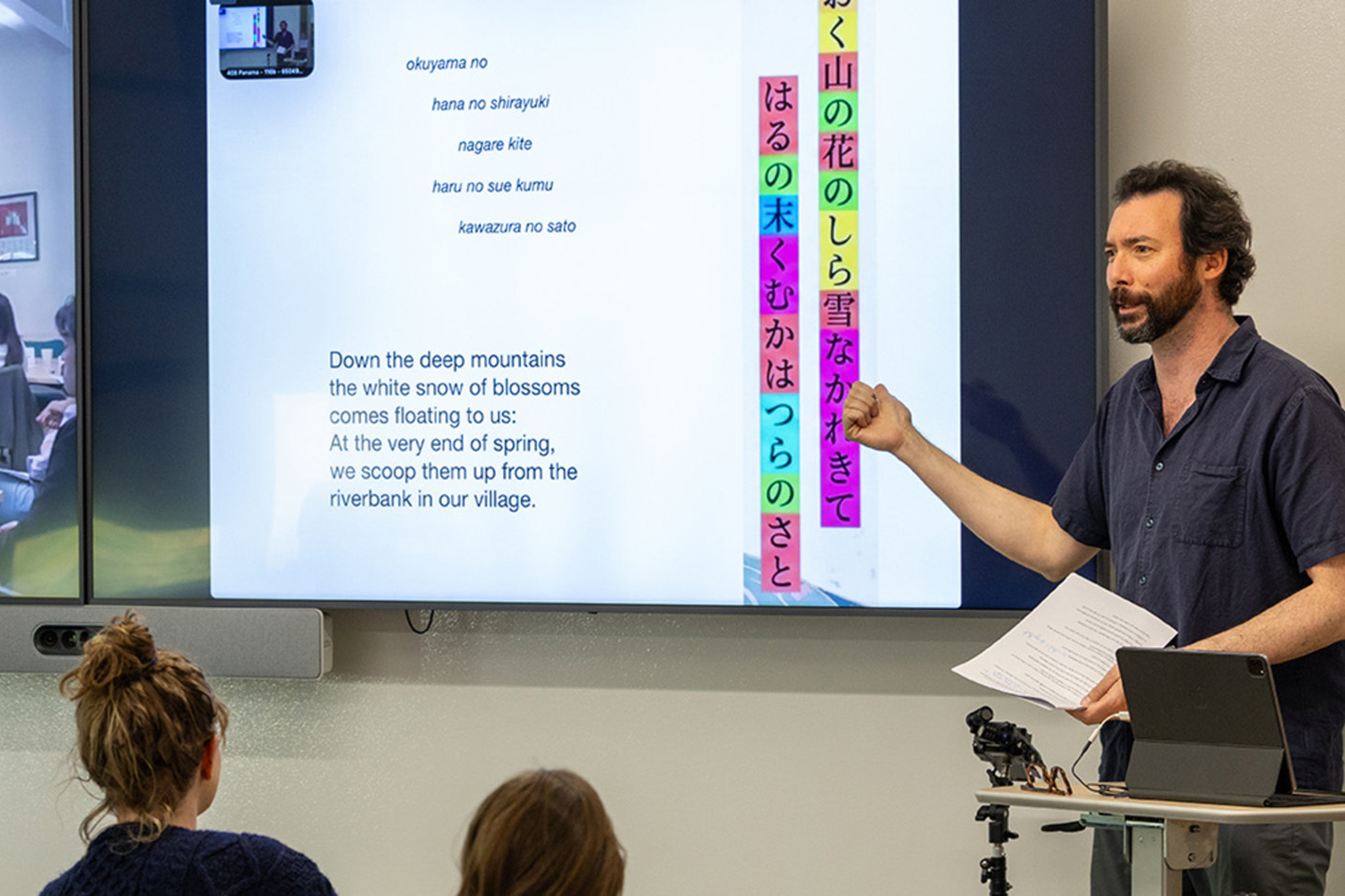 A man gestures while presenting a poem on a screen, with students seated and engaged in a classroom setting.