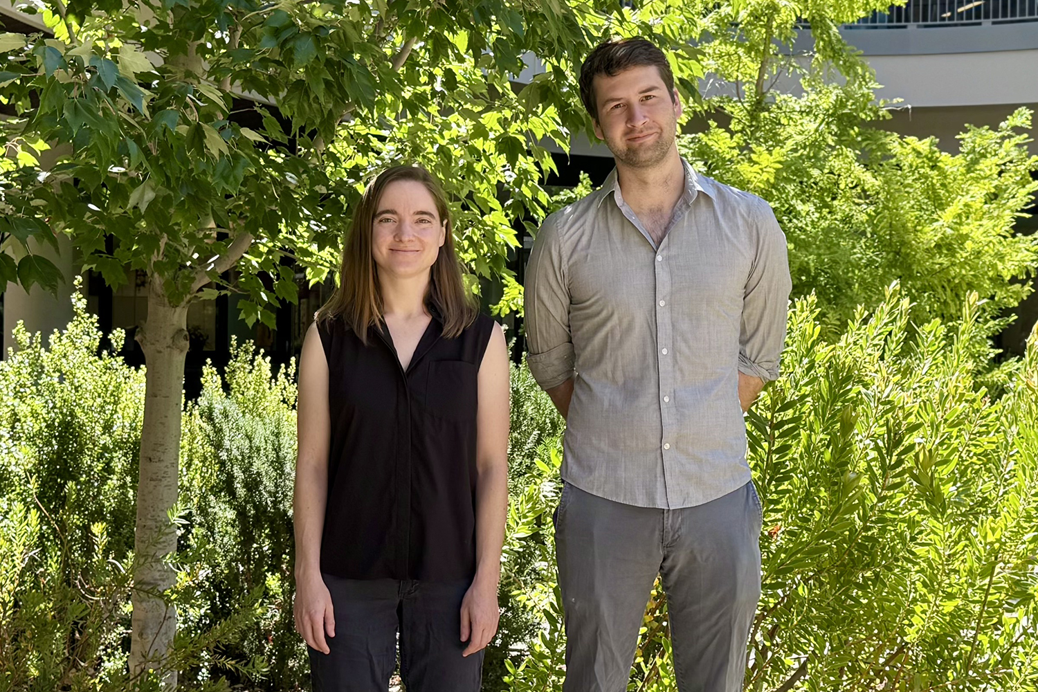 Melissa Gray in a black sleeveless top and Steven Banik in a gray button-down shirt stand outdoors among sunlit trees and shrubs.