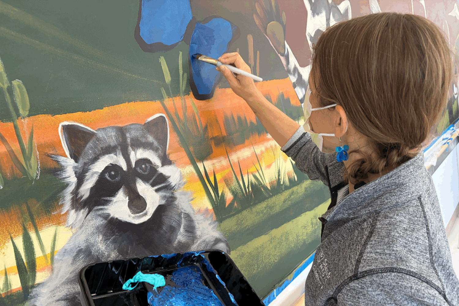 A woman wearing a mask paints a blue butterfly next to a detailed raccoon on a wildlife mural.