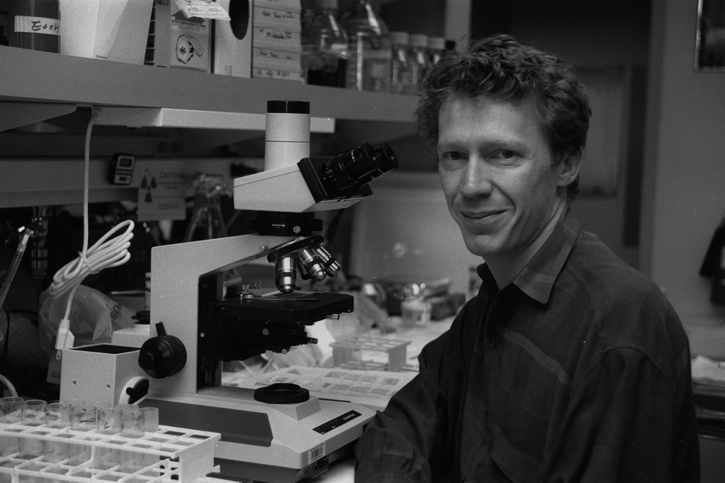 Black-and-white photo of a young Emmanuel Mignot beside a microscope in a laboratory setting.