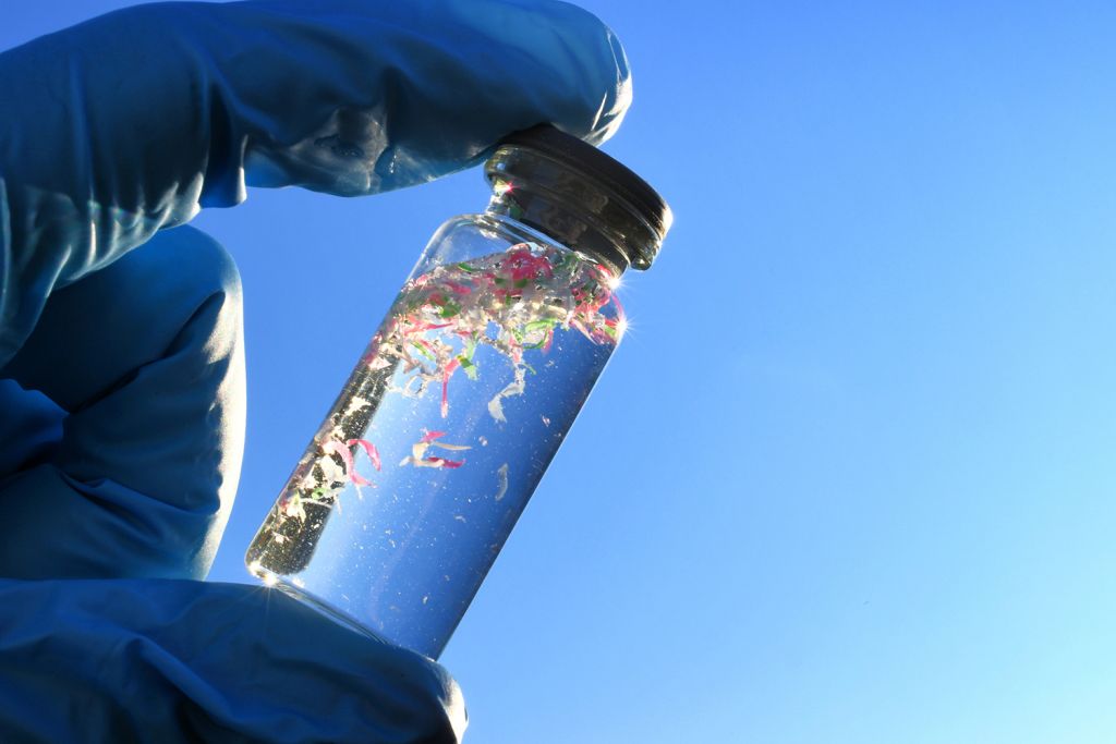 A gloved hand holds a small glass vial of clear liquid with pink, green and white plastic fragments against blue sky.