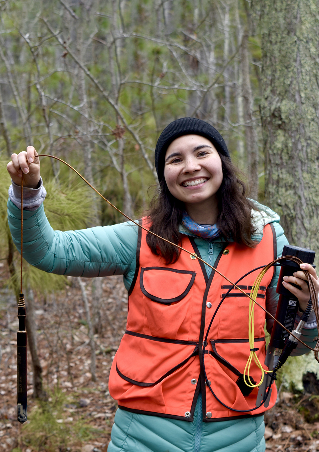 A smiling person in a black beanie and orange vest holds a dangling cable with a weighted probe and a handheld device in a woodland.