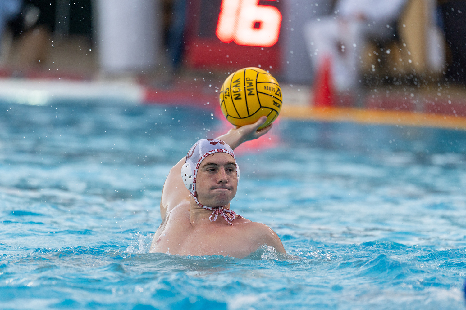 Alex Gheorghe raises a yellow ball in his throwing hand while treading water, with splashes around him and a blurred scoreboard behind.