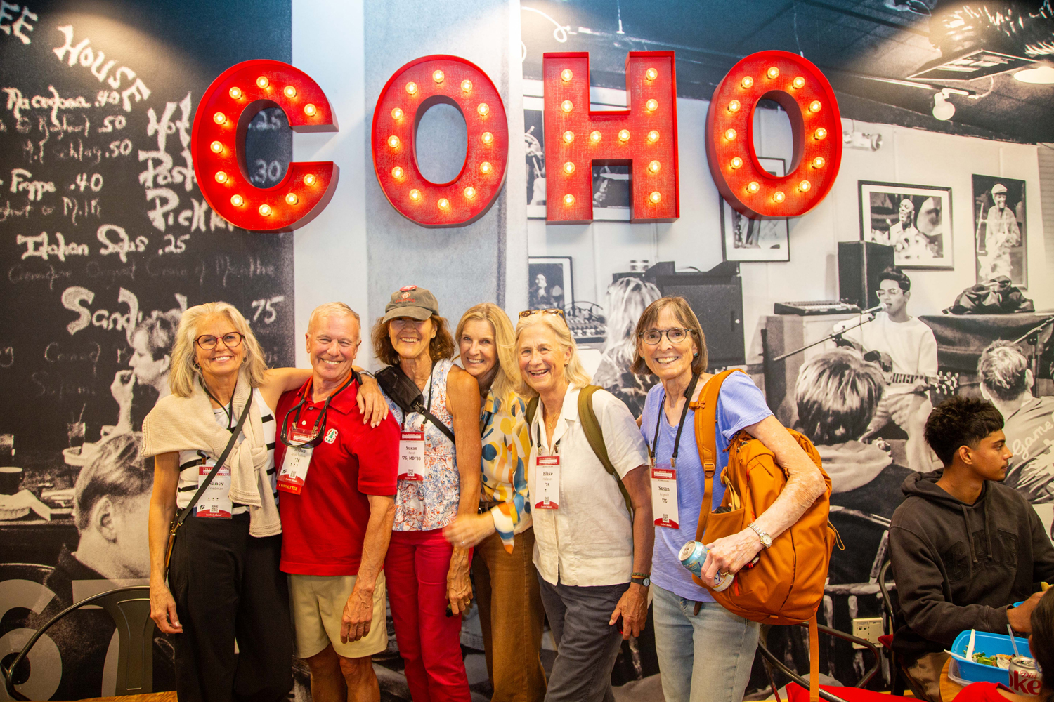 Six smiling alumni pose together under large red marquee letters in a cafe with a black-and-white mural.